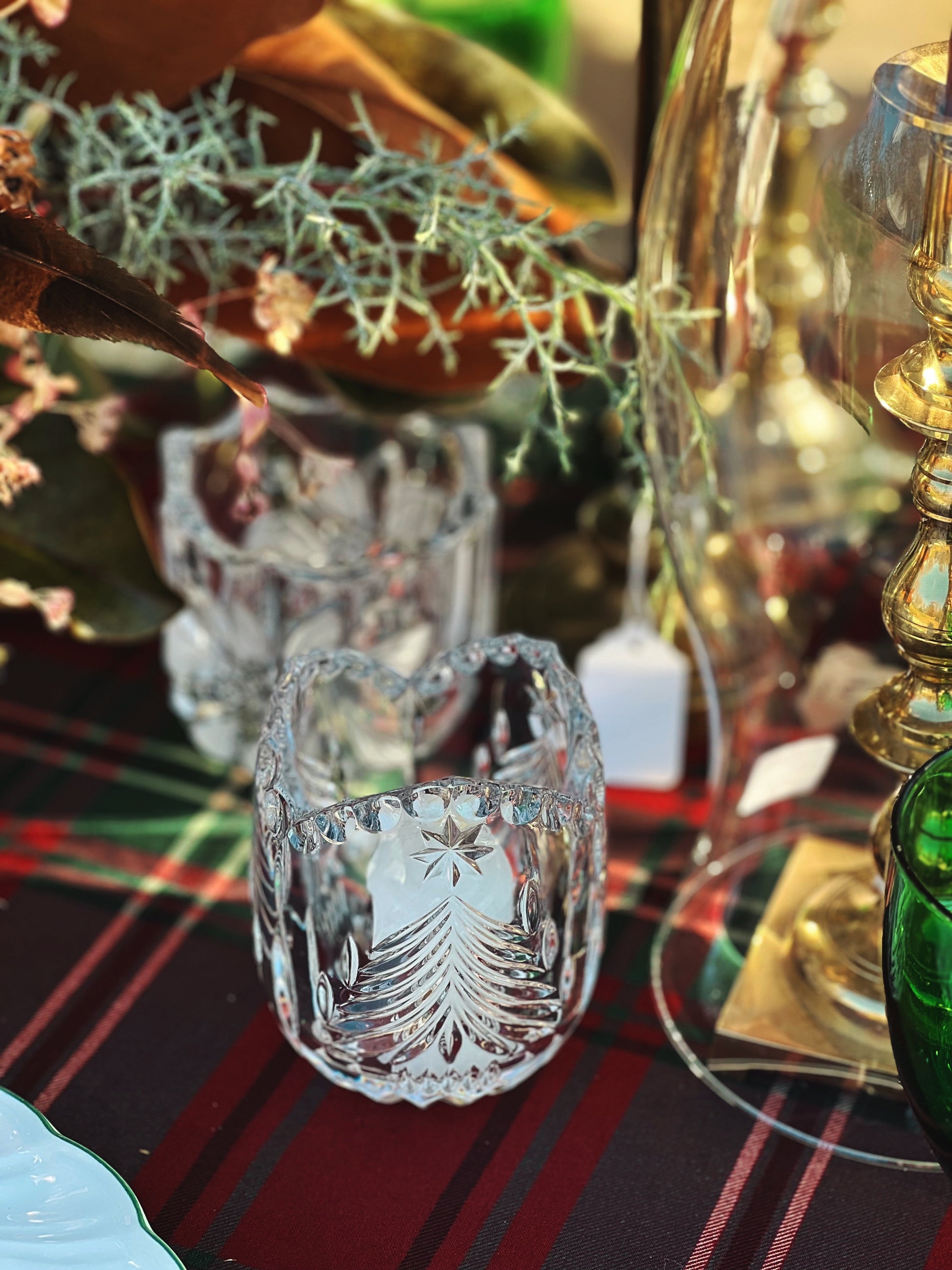 A clear glass votive candleholder with a Christmas tree design, placed on a table with a blurred green and red patterned fabric in the background.