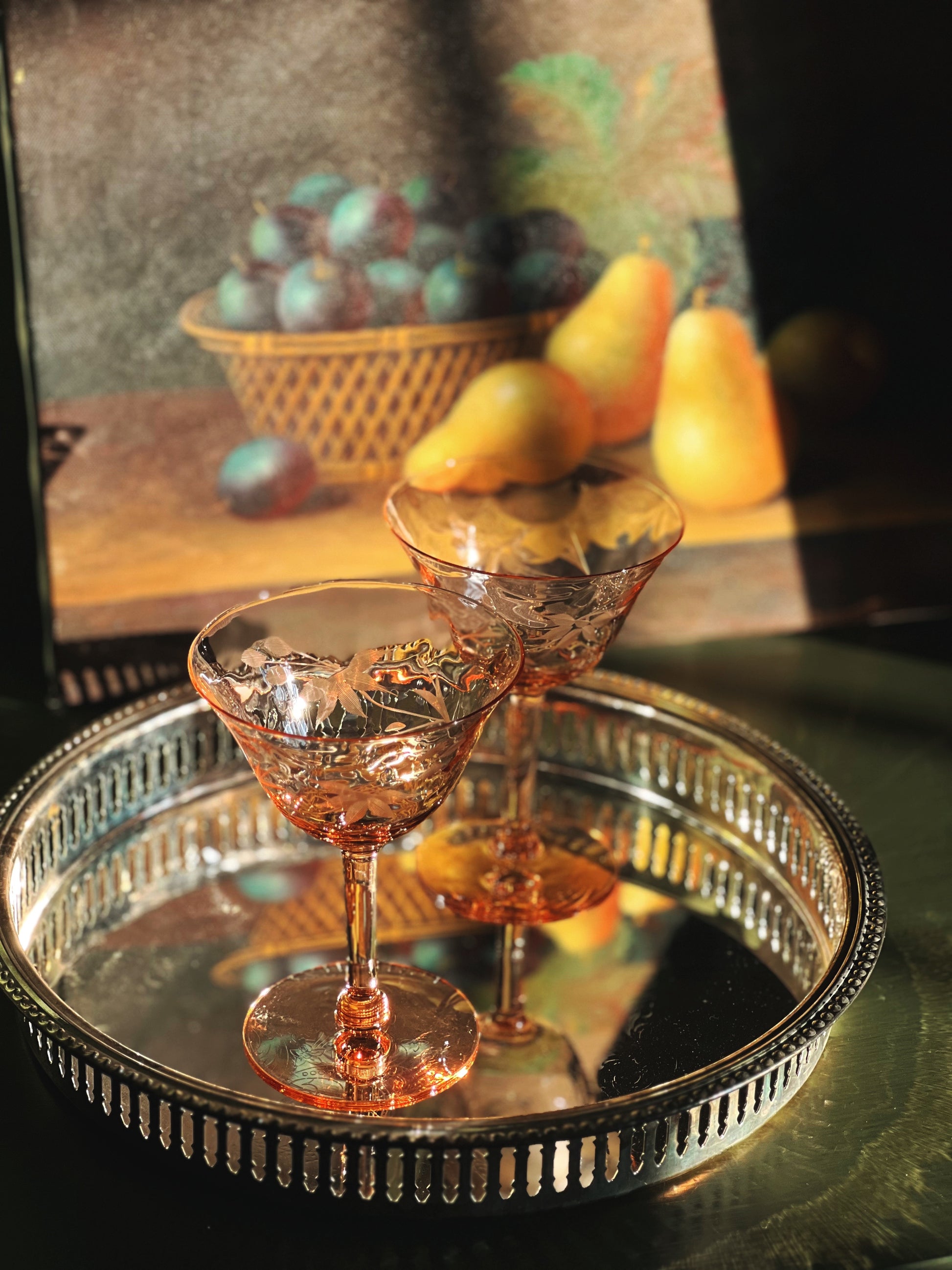Two pink cocktail glasses on a silver tray with a still life painting in the background.