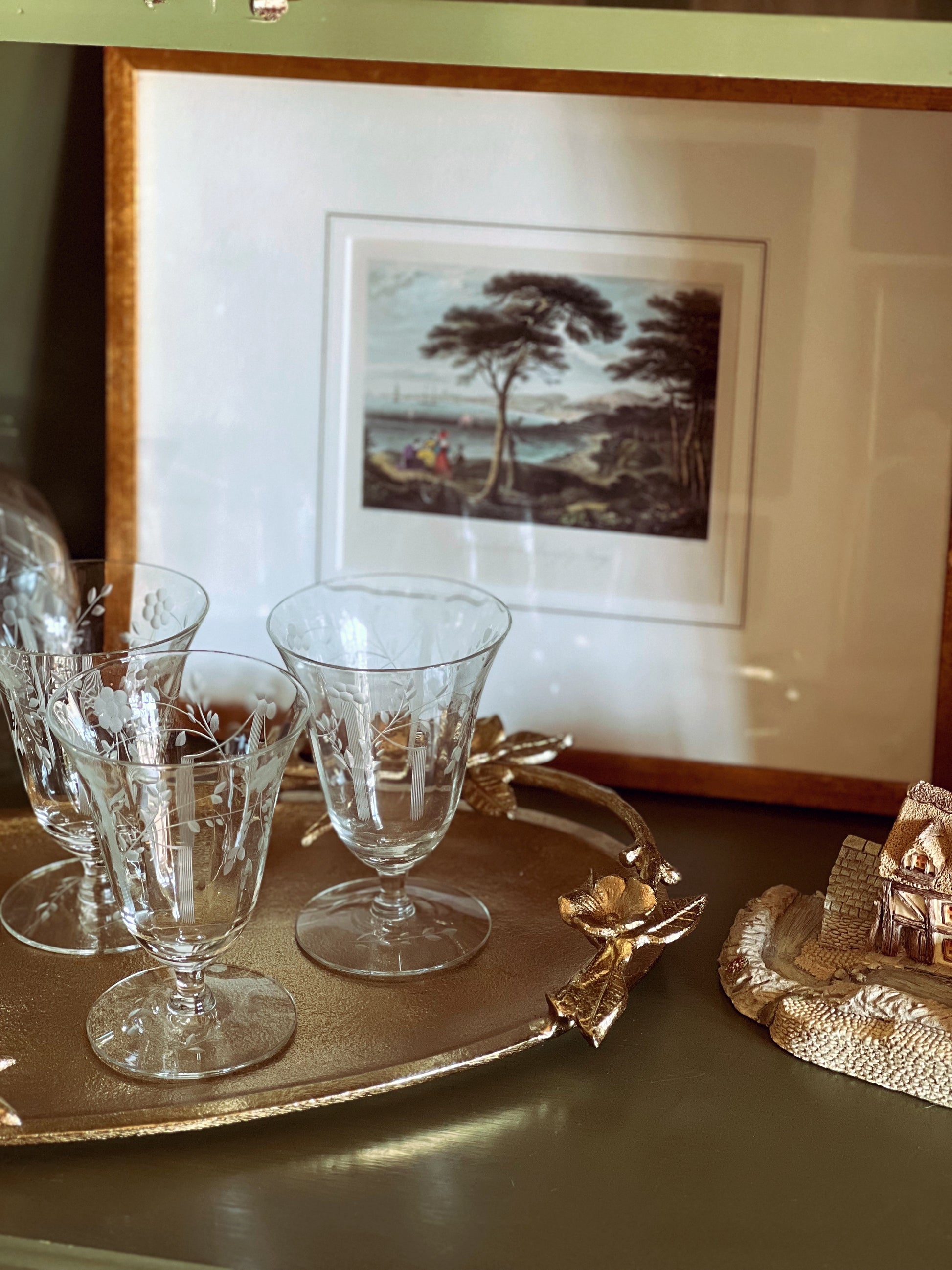 Set of etched glass goblets on a gold tray with a framed painting in the background.