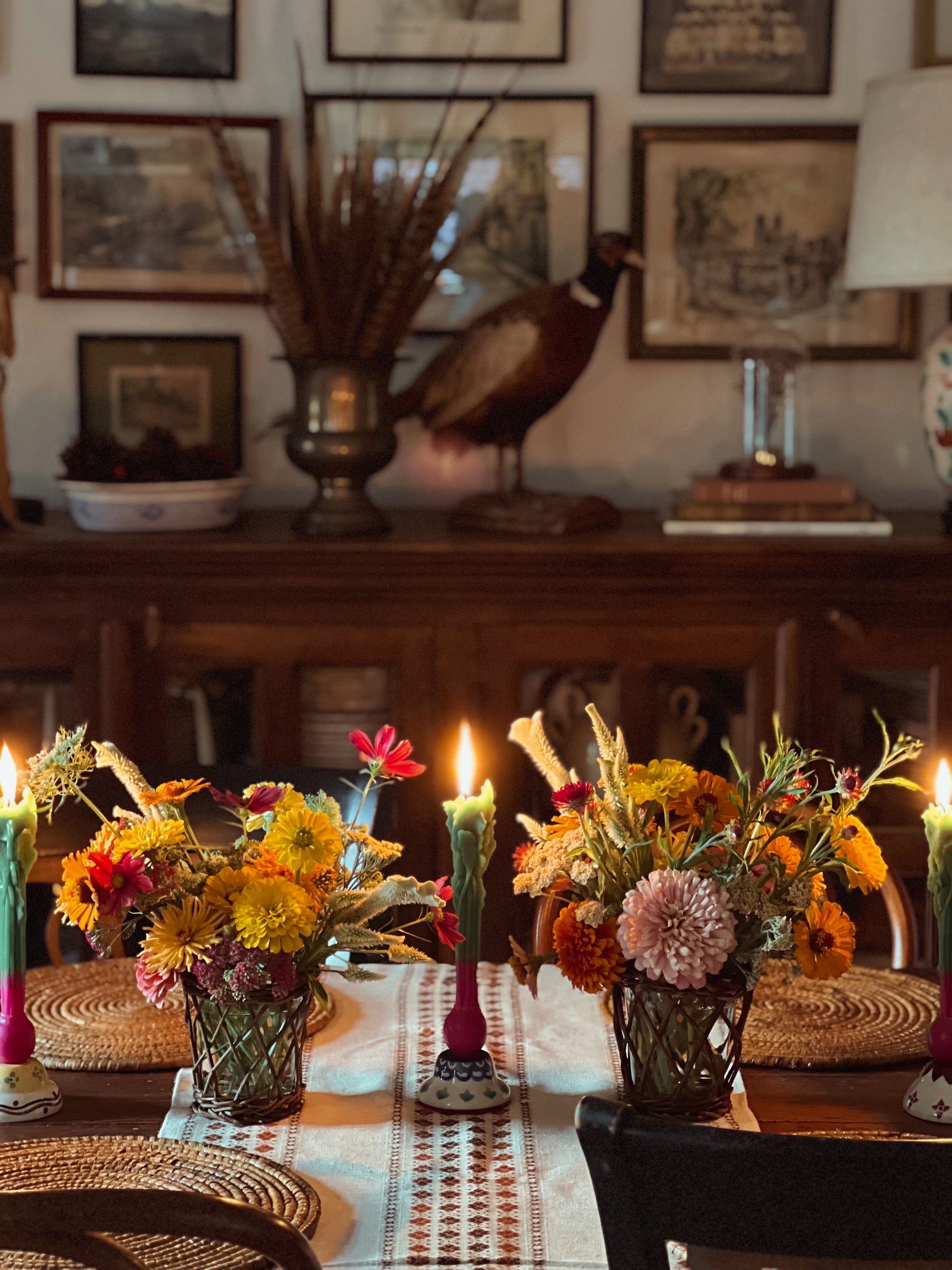 Decorative table setting with flowers, candles, and a peacock statue in a room with framed pictures on the wall.