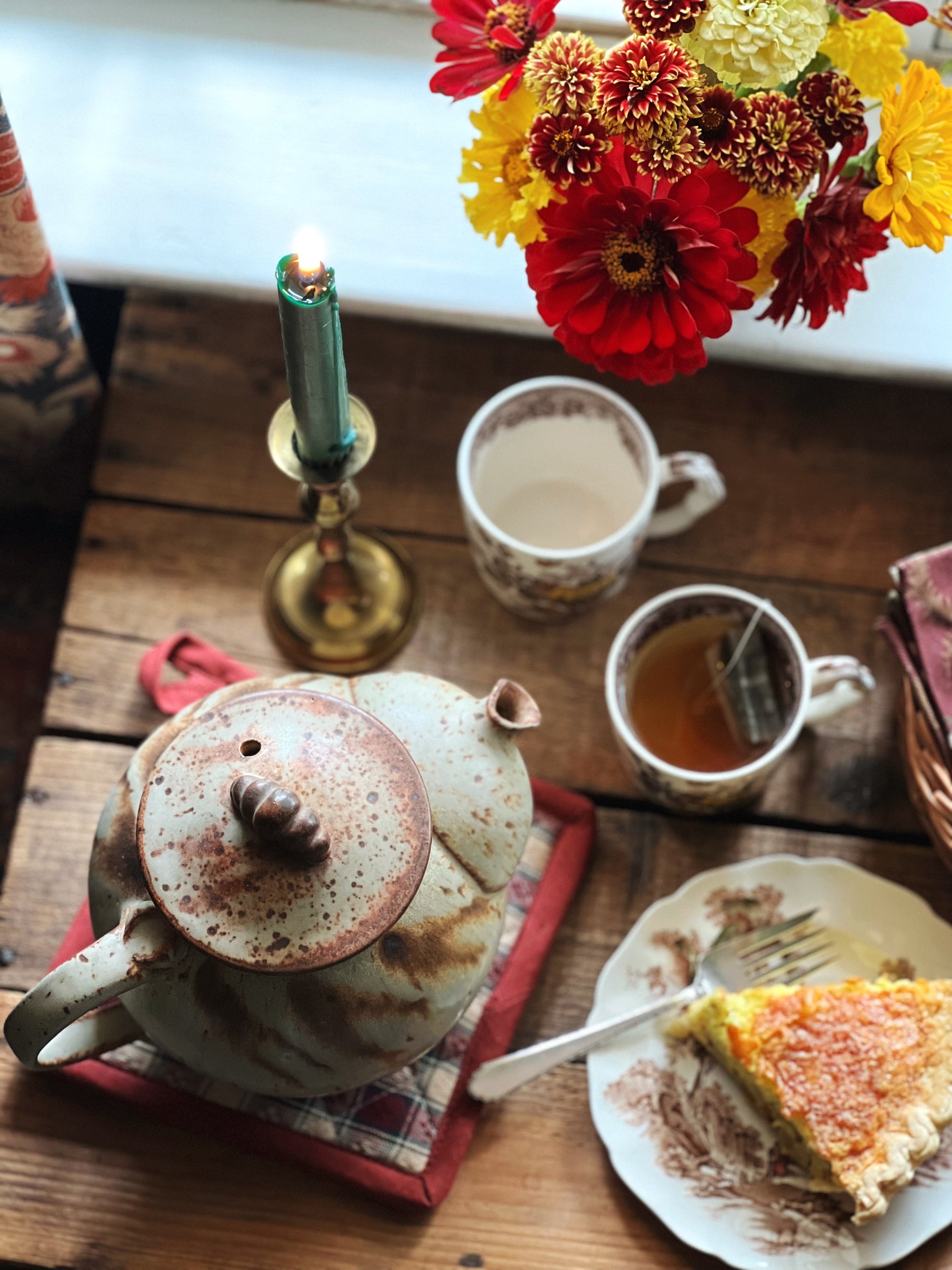 A vintage stoneware teapot with a round, embossed body in a brown and green color scheme, sitting on a red cloth next to a cup of tea and a slice of cake.