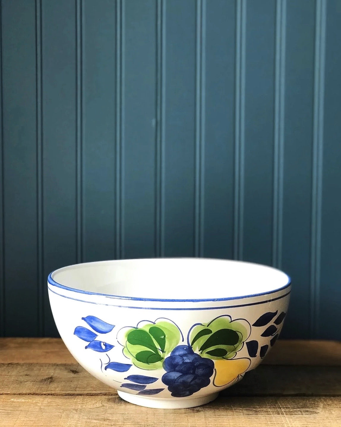A hand-painted, footed ceramic serving bowl with blue and white floral patterns on a green background, placed on a wooden surface against a blue paneled background.