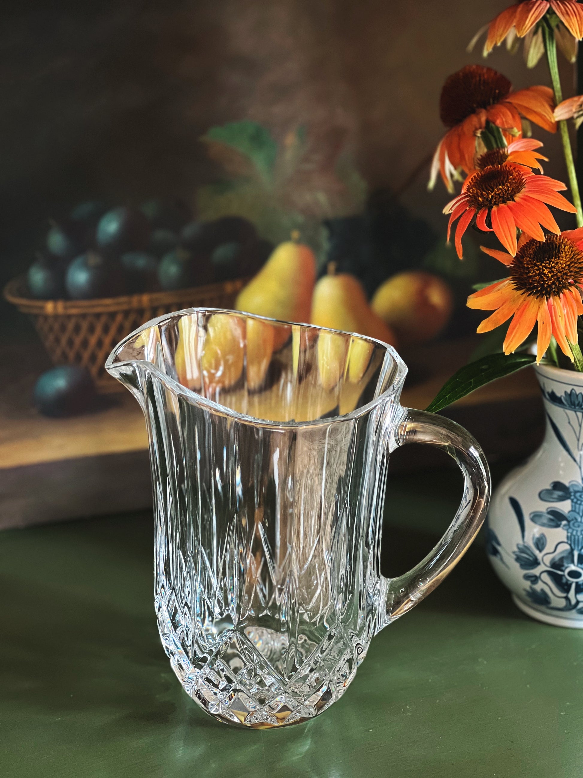 Clear glass pitcher on a table with fruits and flowers in the background