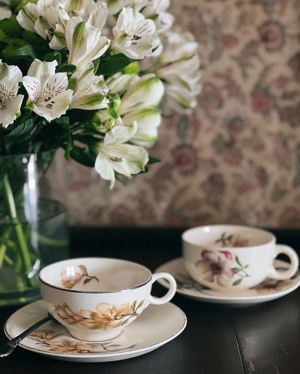 Teacups with floral design on saucers next to a vase of white flowers against a patterned wall.