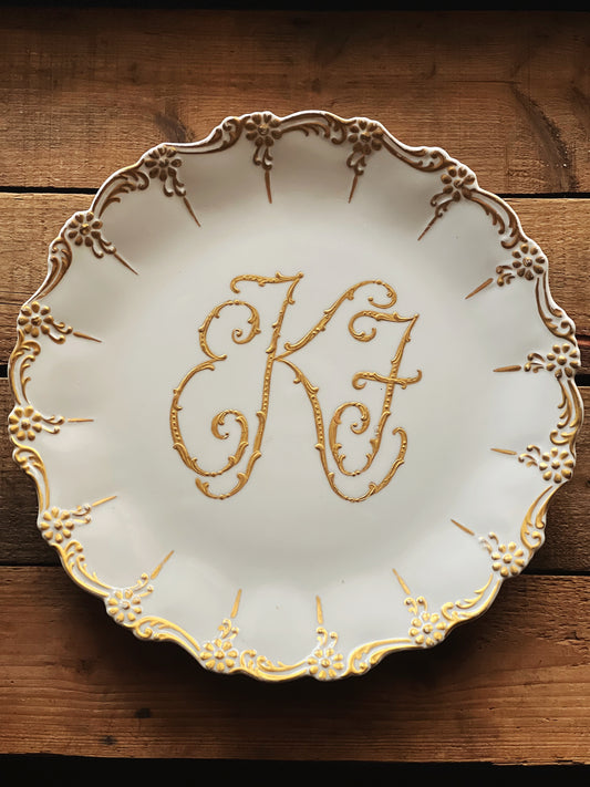 white platter with a gold monogram and elaborate rim design 