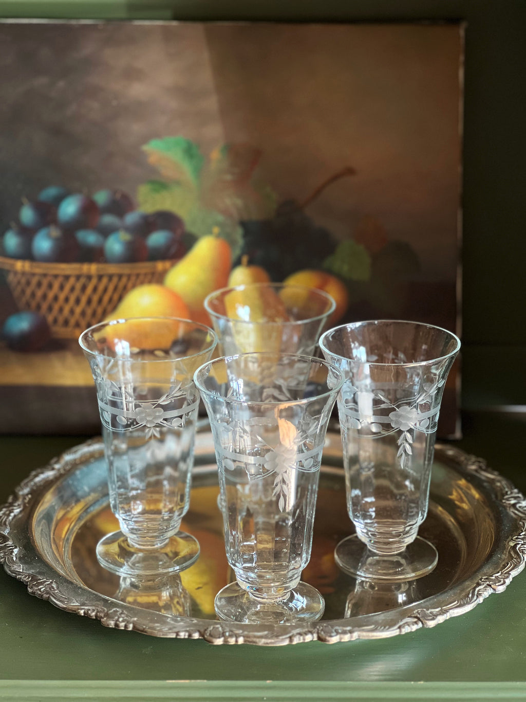 Set of six etched glass tumblers on a silver tray with a fruit painting in the background.
