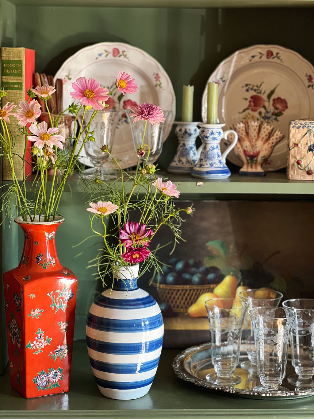 Decorative shelf with floral arrangements, vases, and tableware against a green wall.