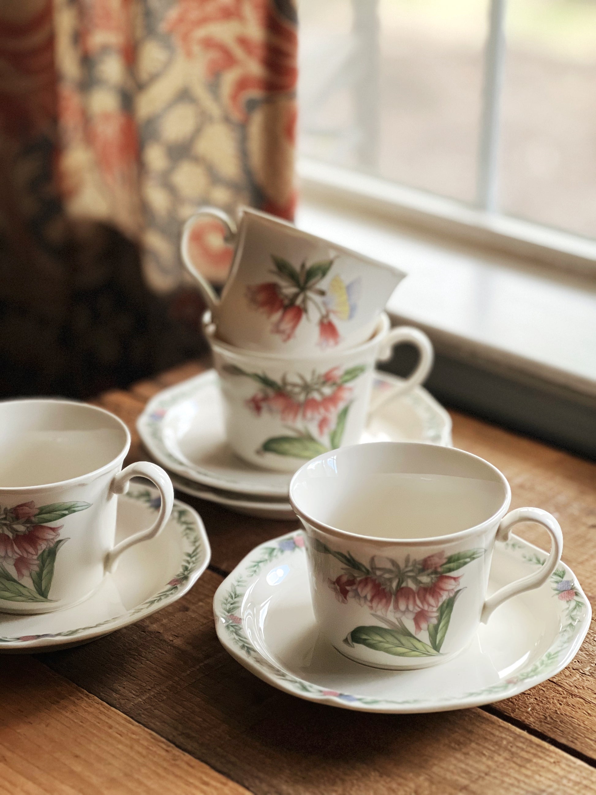 Set of floral teacups and saucers on a wooden table near a window.