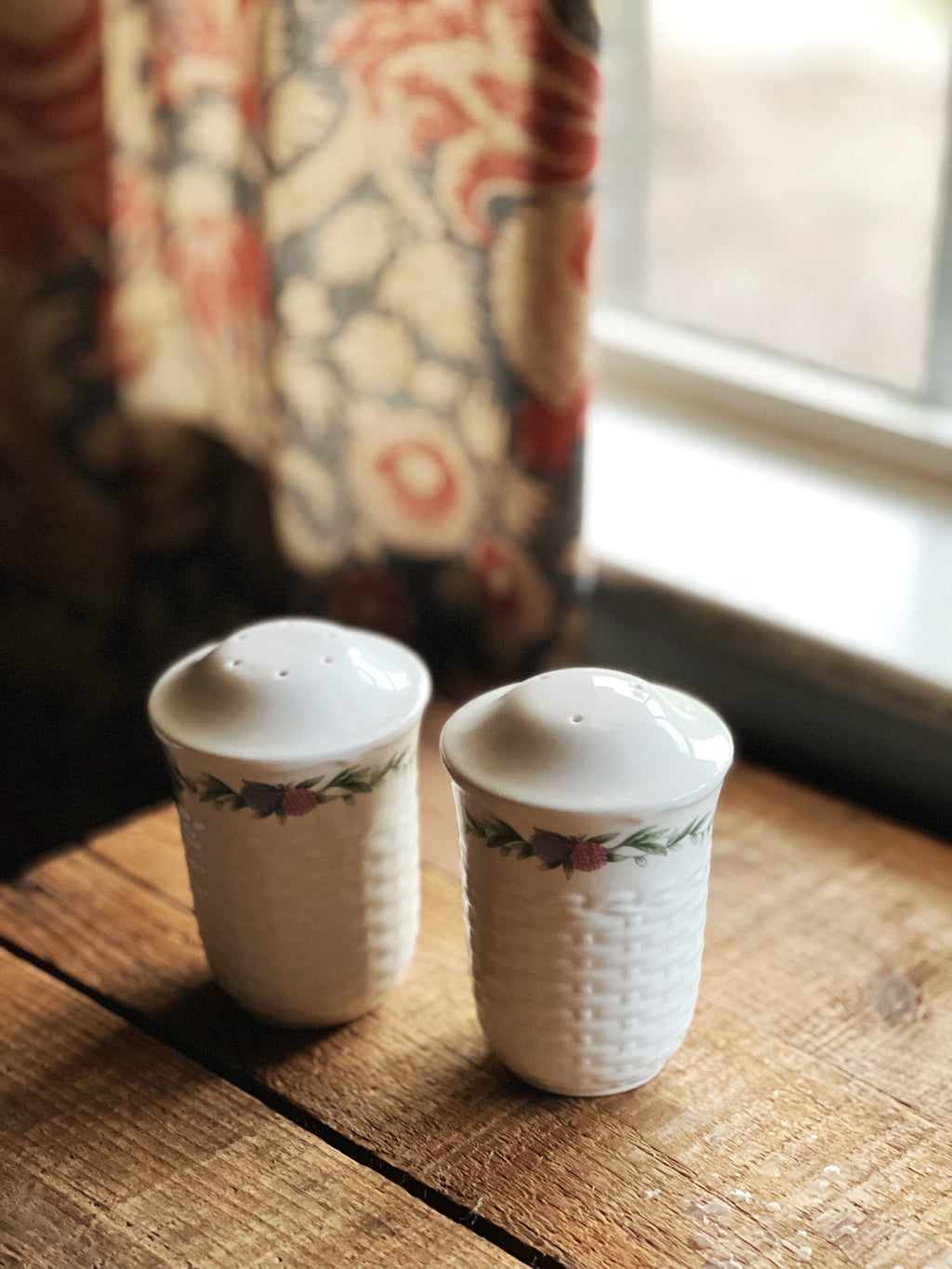 Two ceramic salt and pepper shakers on a wooden surface with a floral curtain in the background.