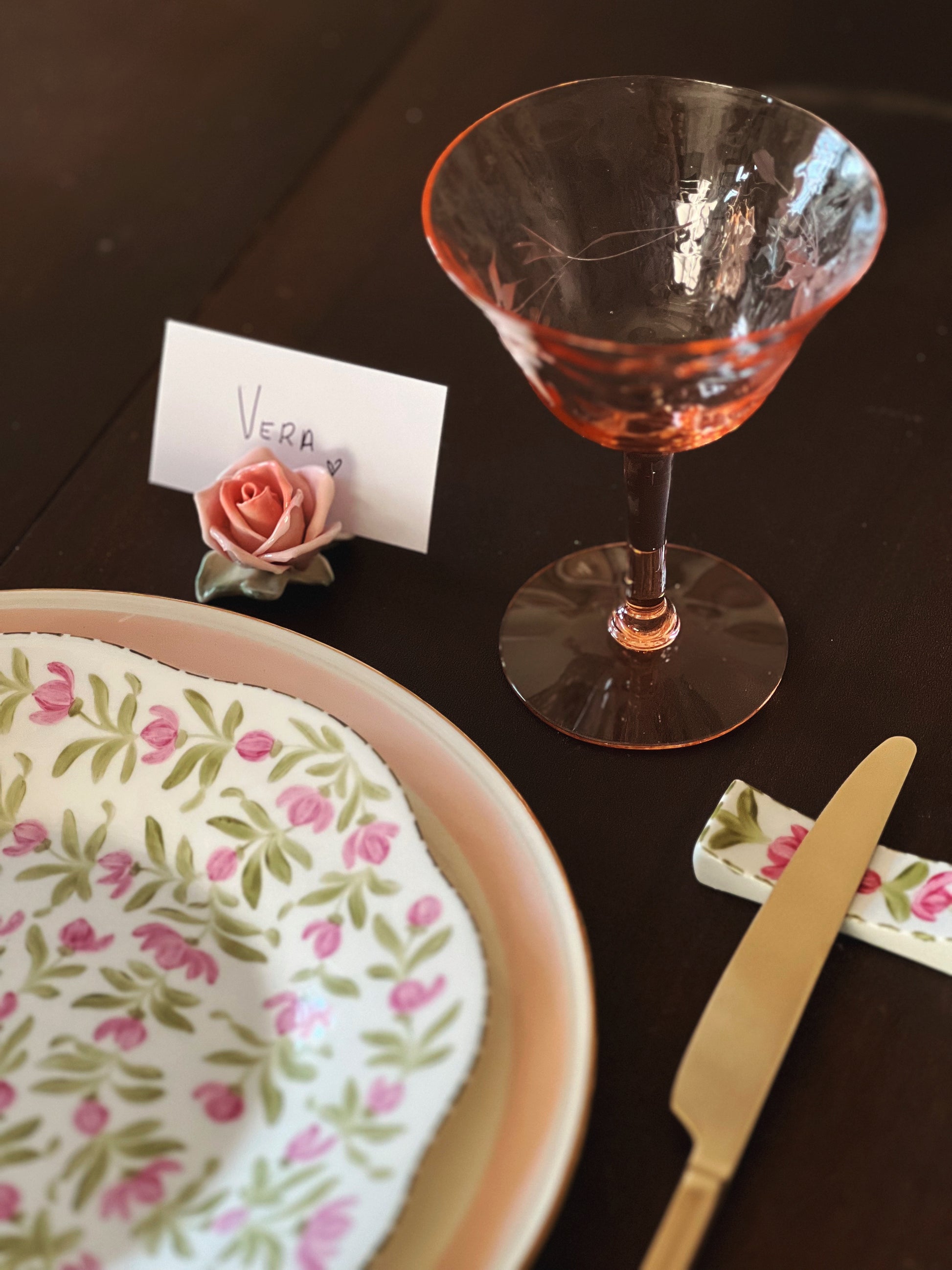 Table setting with a floral plate, pink glass, and decorative elements on a dark surface.