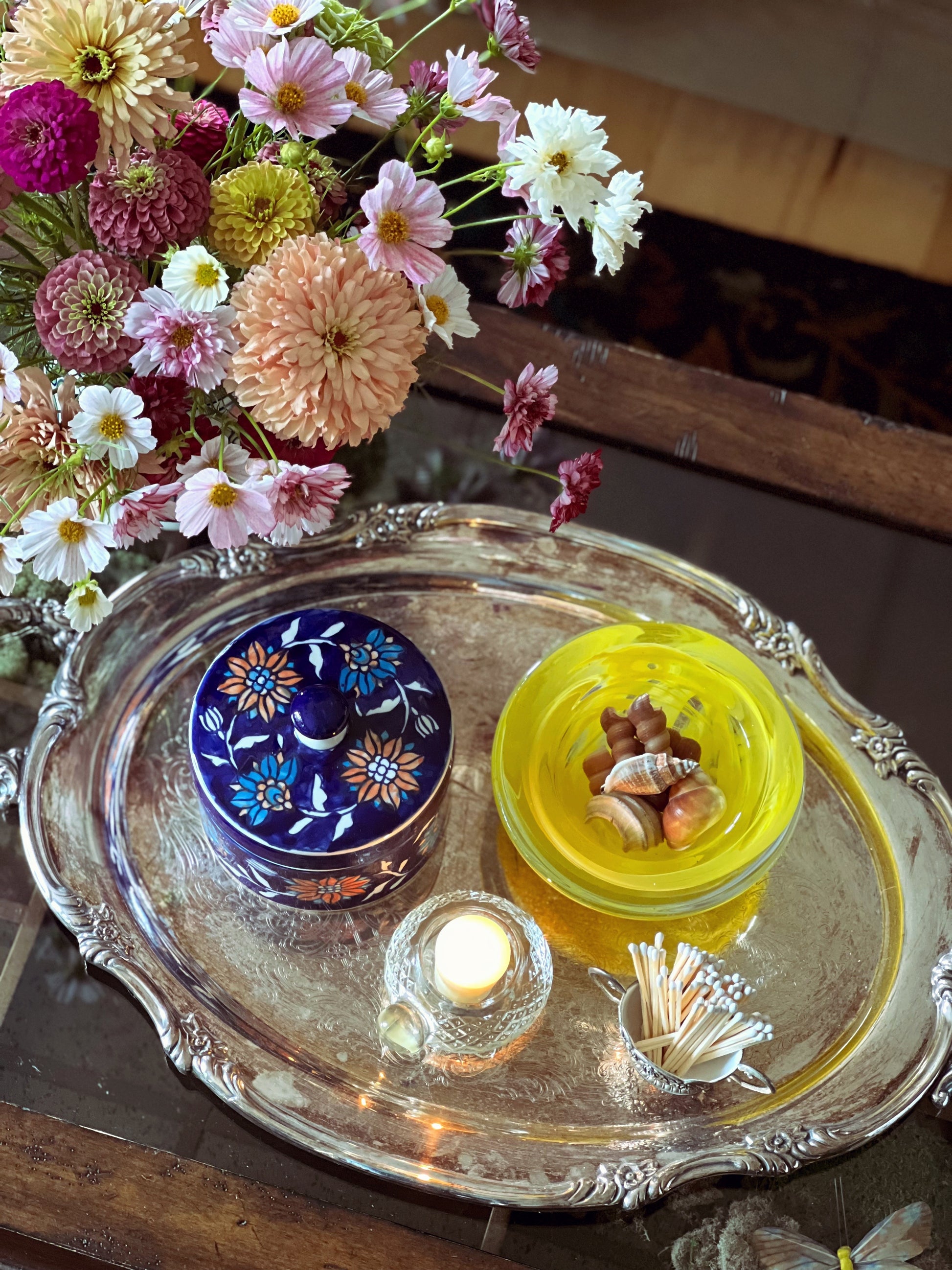 Decorative tray with floral arrangement, candles, and matchbox on a wooden surface.