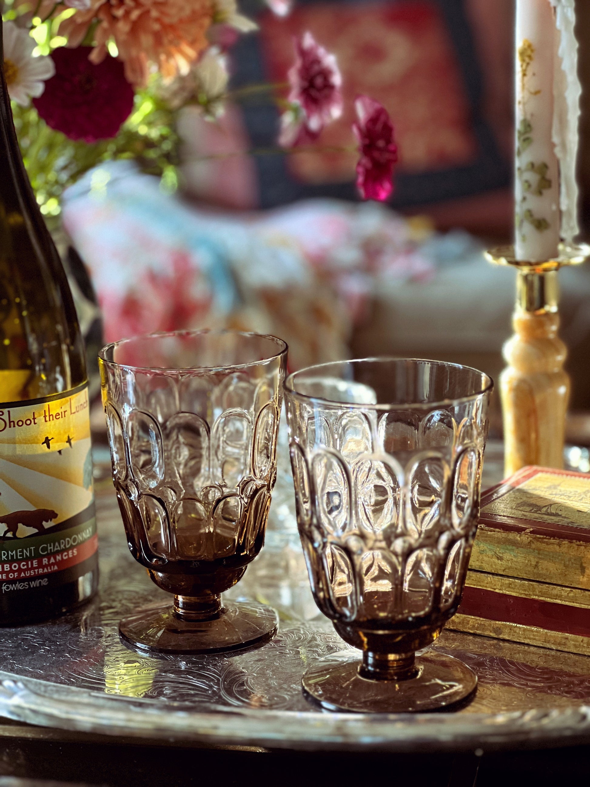Two ornate glass goblets on a table with a bottle and books in the background.