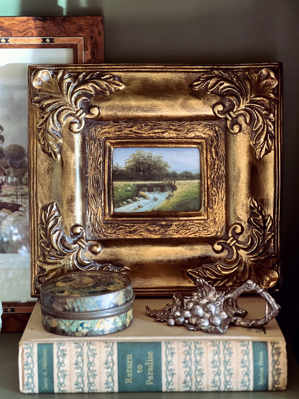 Decorative setup with ornate gold frame, small painting, books, and decorative items.