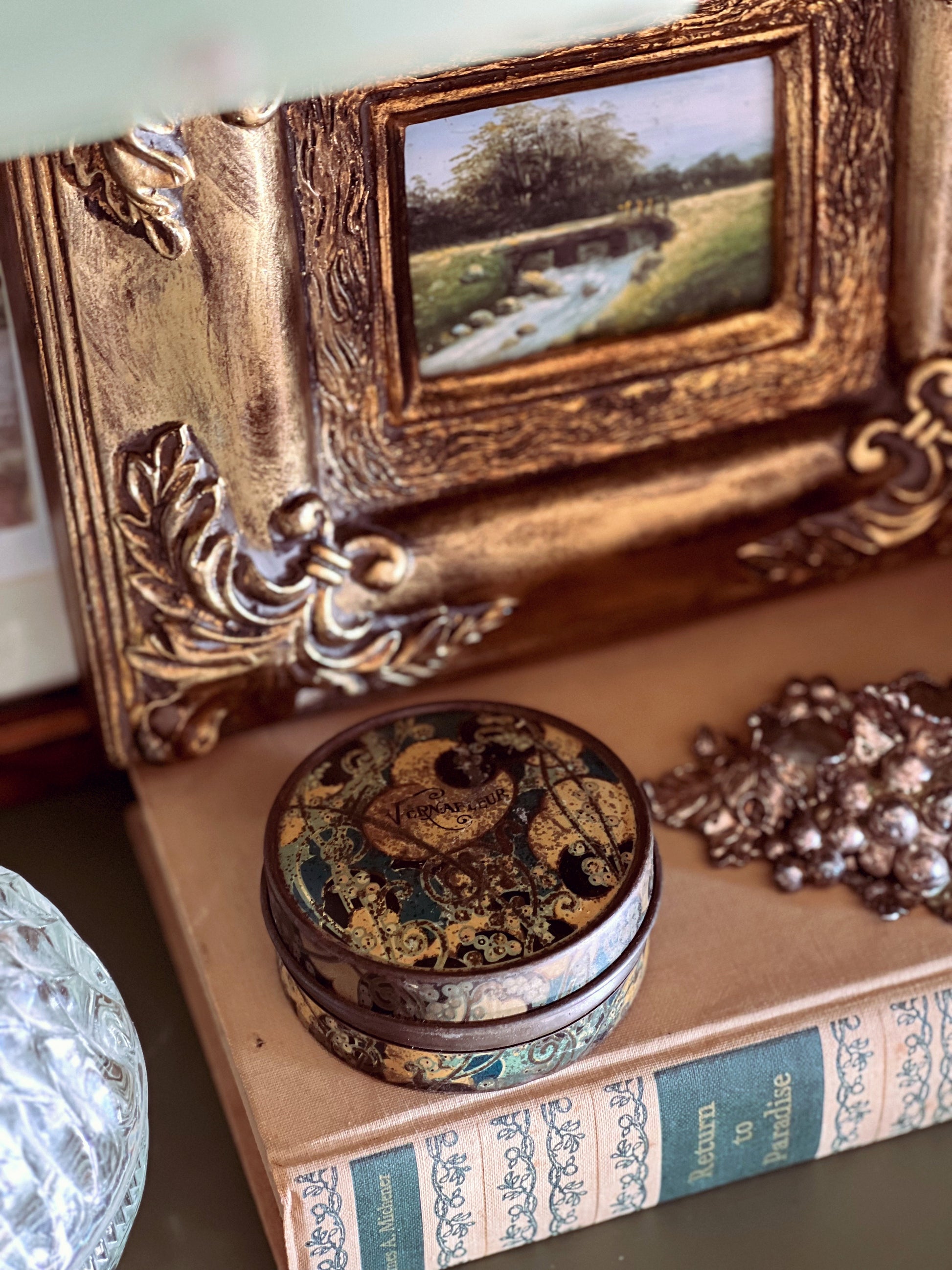 Decorative gold frame with a small painting, ornate box, and jewelry on an old book.