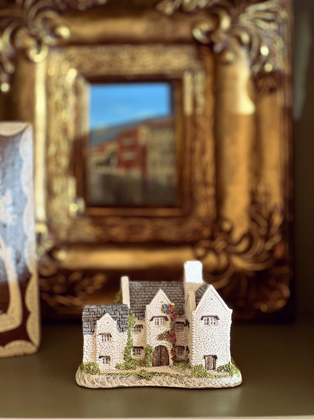 Small model of a house with decorative elements in front of an ornate gold frame.