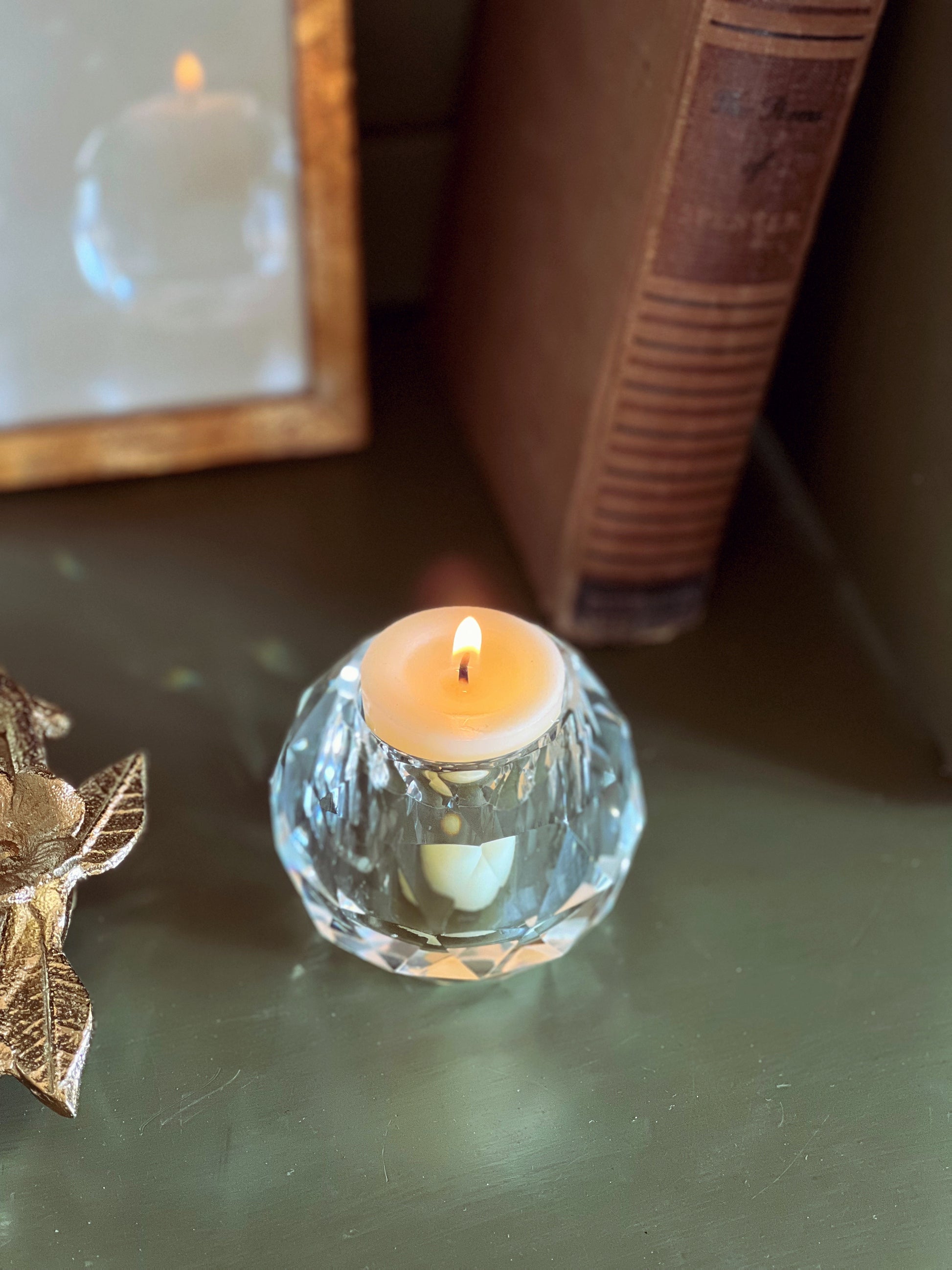 Small lit candle in a glass holder on a reflective surface with a book and decorative item in the background.