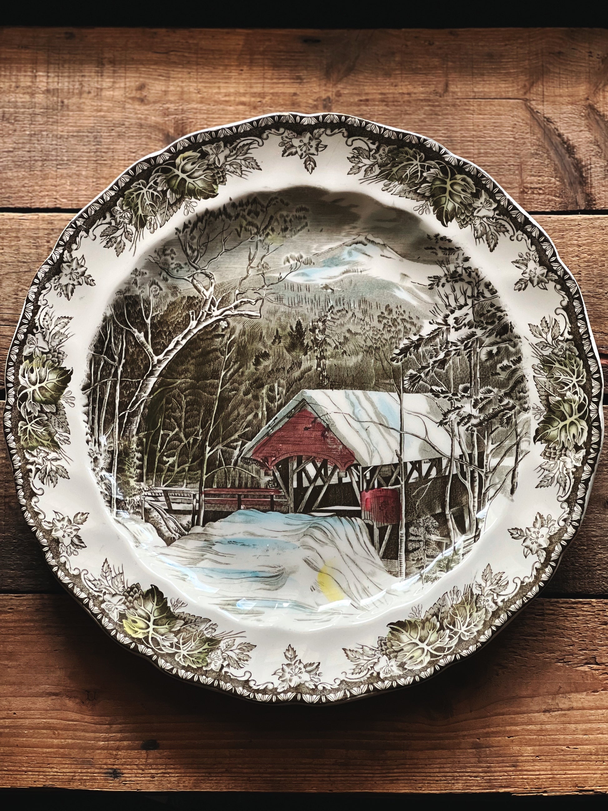 Decorative plate with a winter scene featuring a covered bridge on a wooden surface
