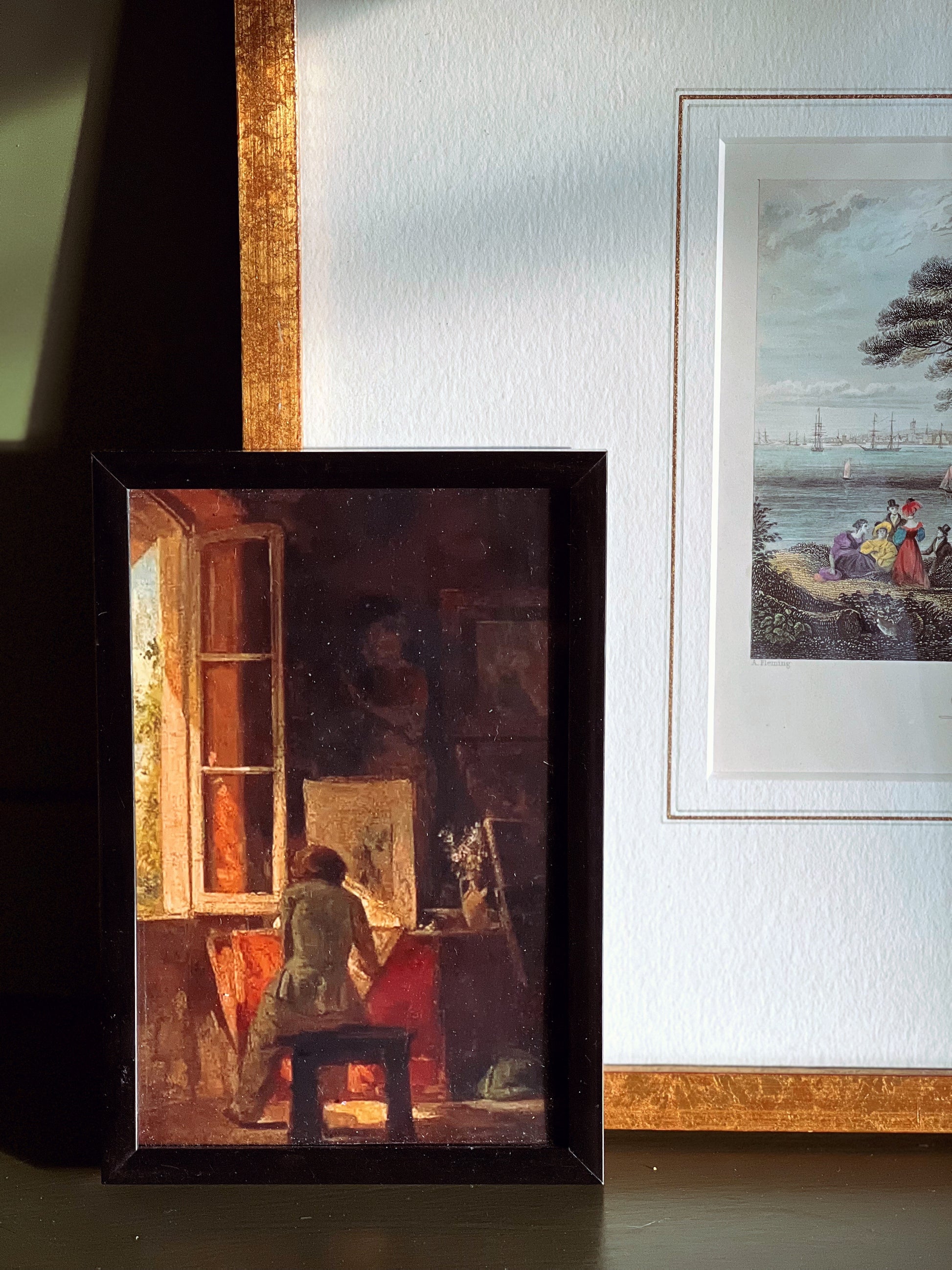 Framed painting of a woman painting a landscape on a wall with another painting in the background.