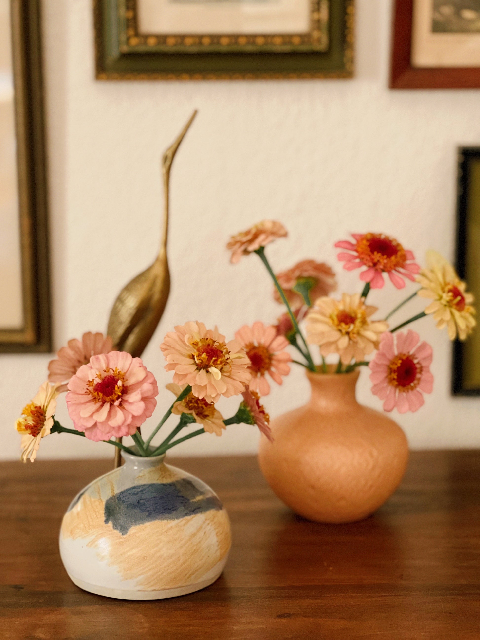 Two ceramic vases with flowers on a wooden surface, framed pictures in the background