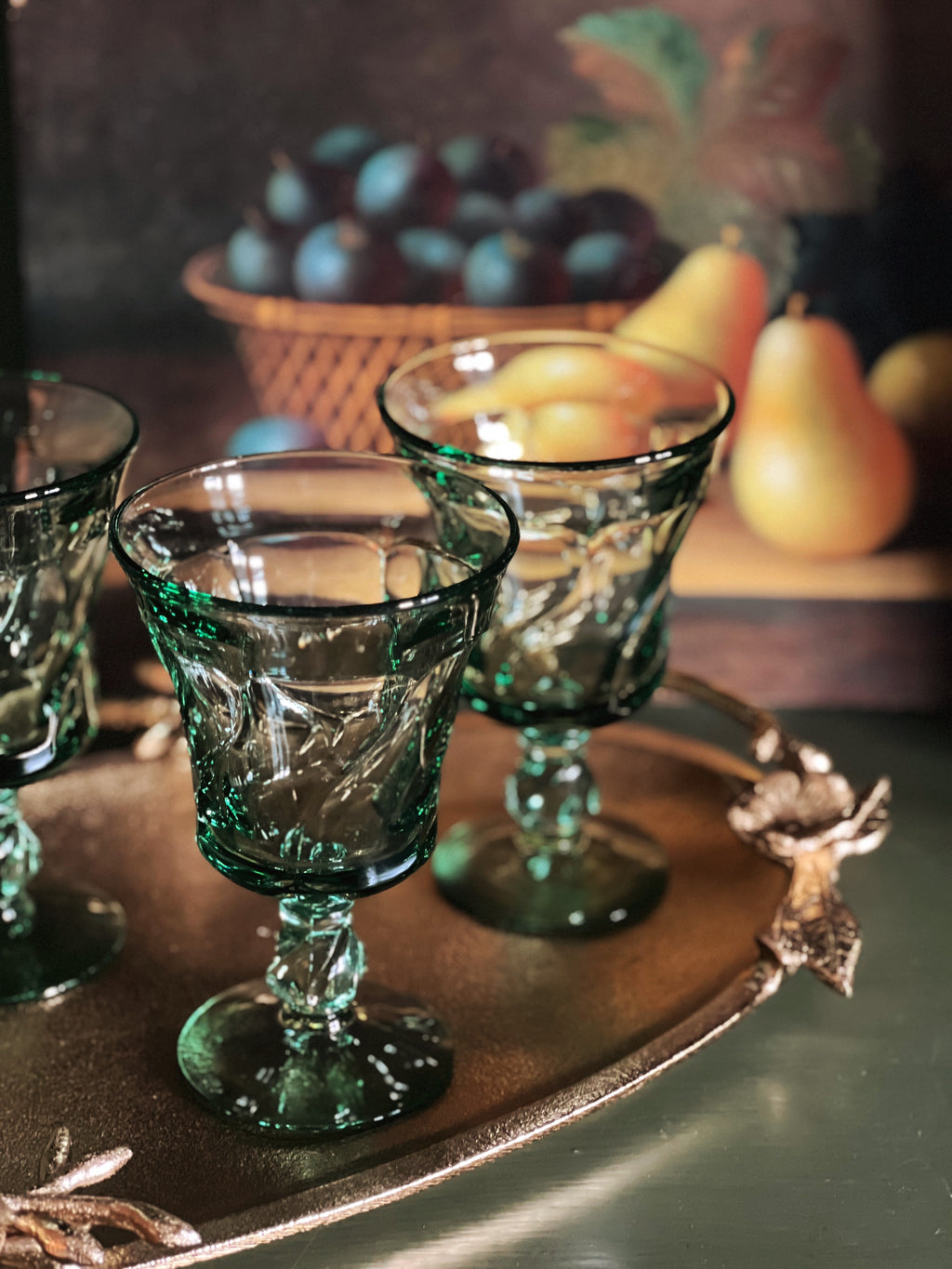 green wine glasses on a gold tray with a still life painting in the background
