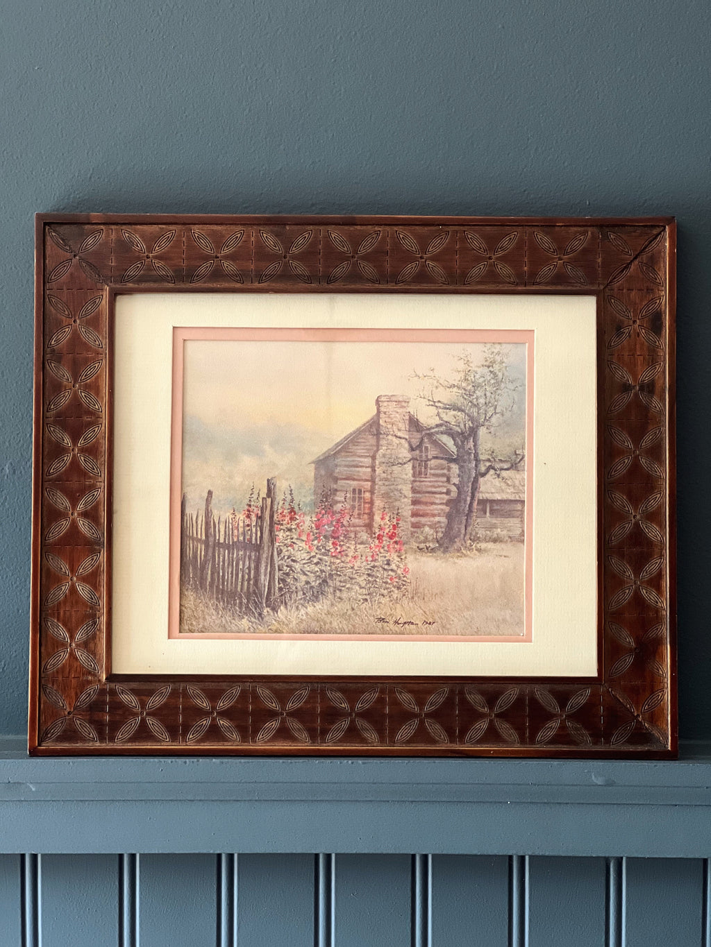 Framed artwork of a rustic scene with a cabin on a blue wall.