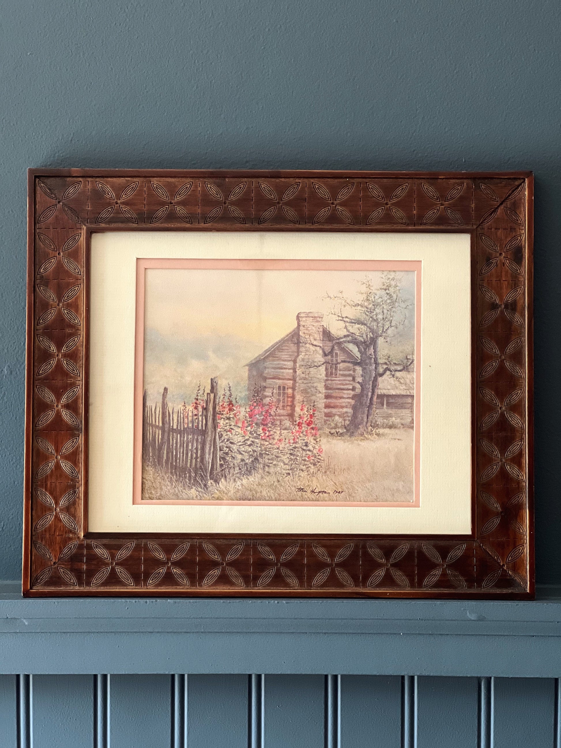 Framed artwork of a rustic scene with a cabin on a blue wall.