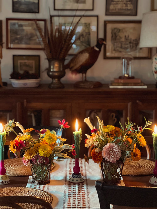 Decorative table setting with flowers, candles, and a peacock statue in a room with framed pictures on the wall.