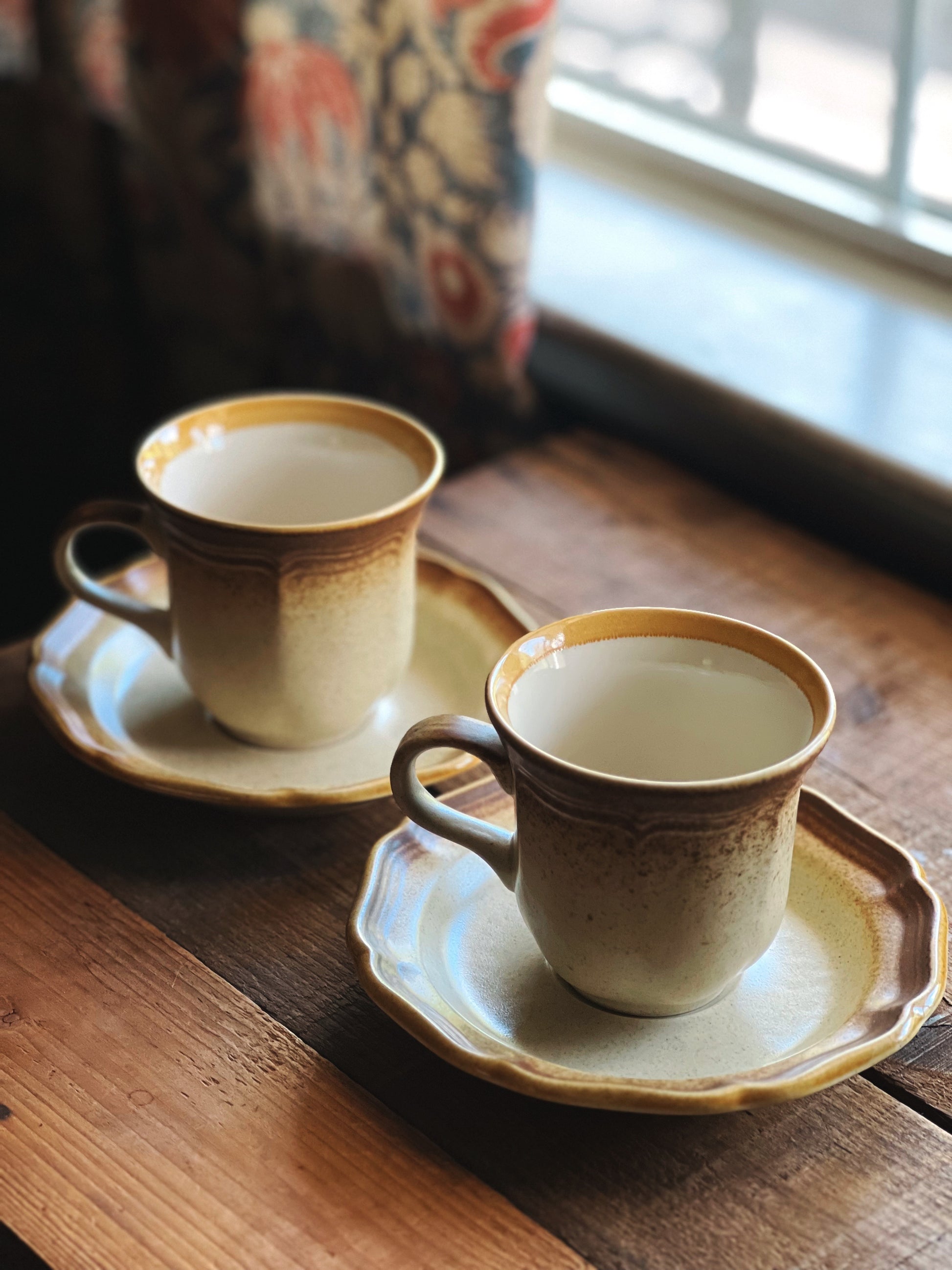 Two ceramic cups with saucers on a wooden surface near a window.