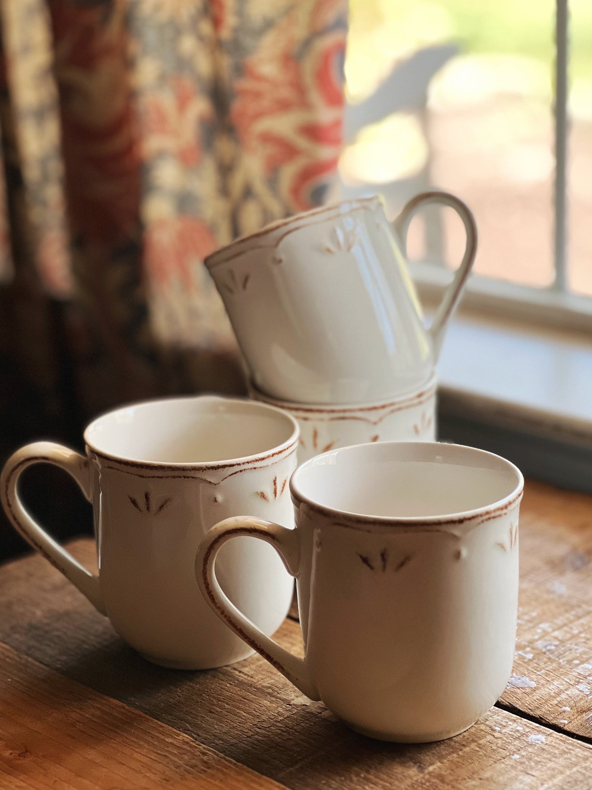 Three ceramic mugs on a wooden surface with a window and floral curtain in the background.