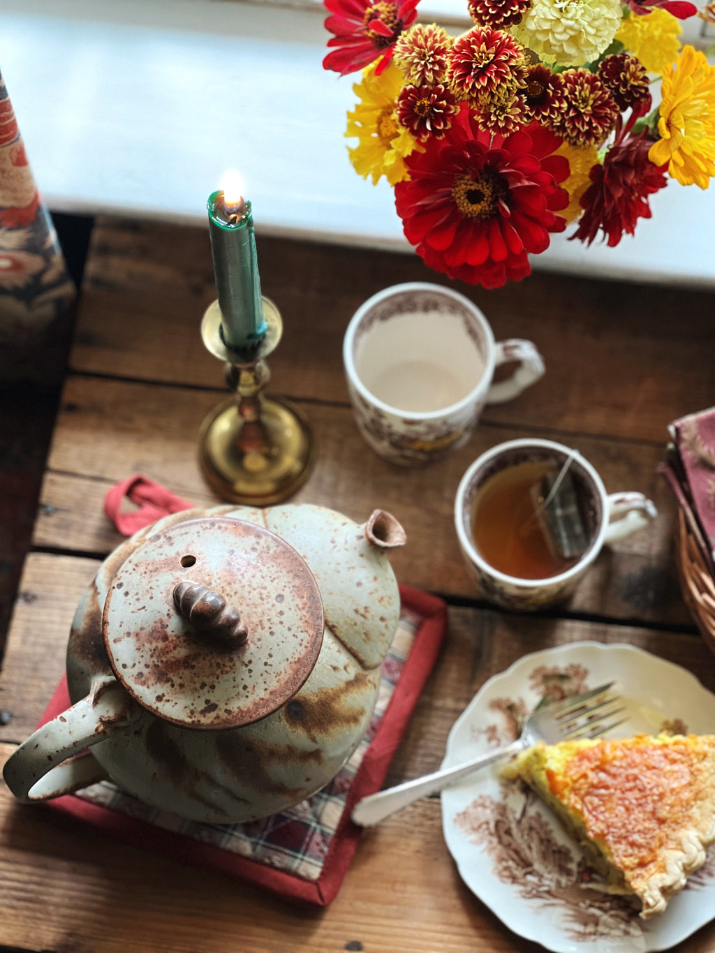A vintage stoneware teapot with a round, embossed body in a brown and green color scheme, sitting on a red cloth next to a cup of tea and a slice of cake.