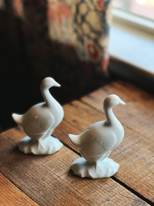 Two white ceramic goose figurines on a wooden surface with a blurred background