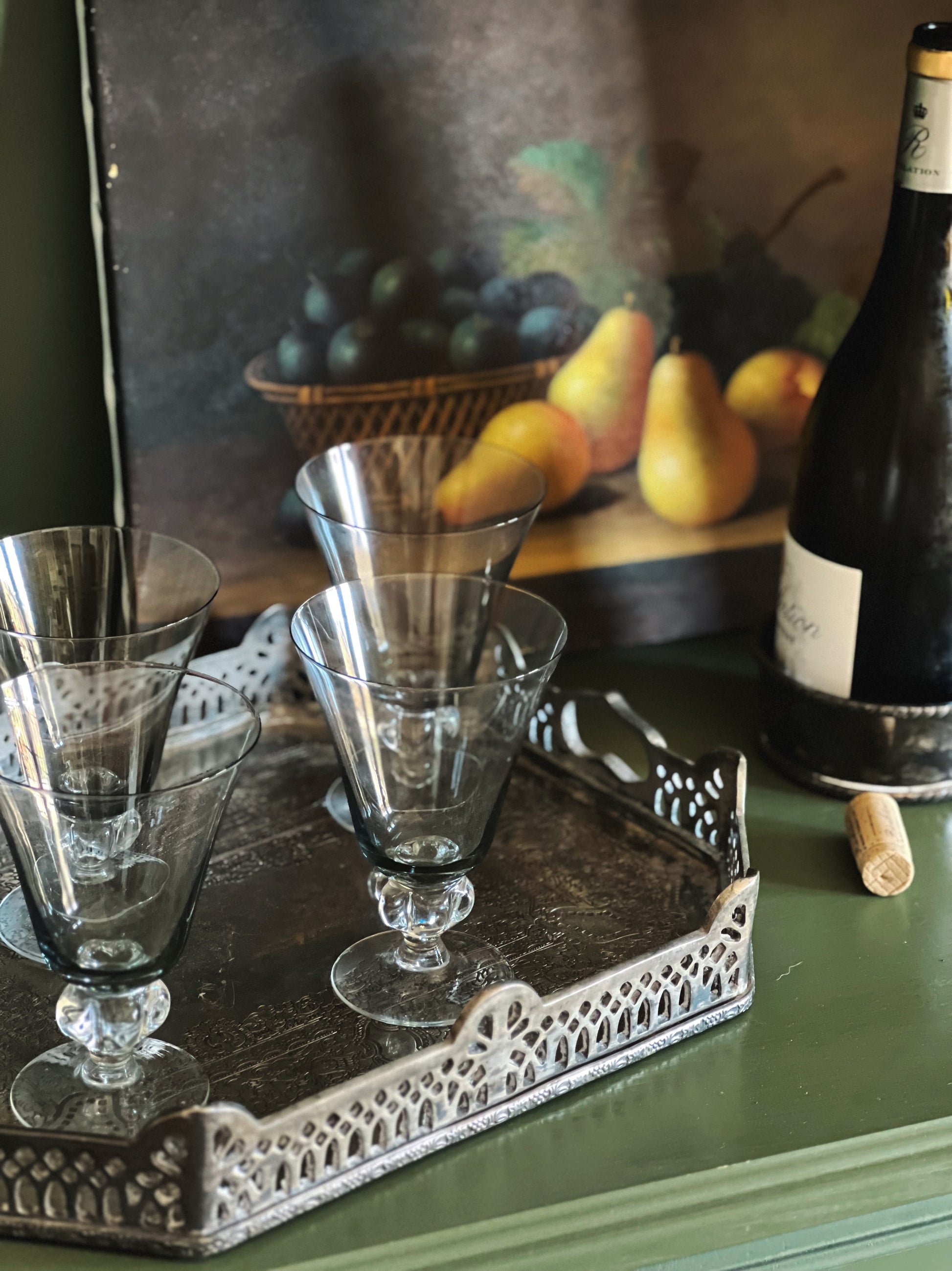 A set of two smoky gray glass wine glasses with a decorative stem design, displayed on a table with a fruit painting in the background and a bottle of wine in the foreground.