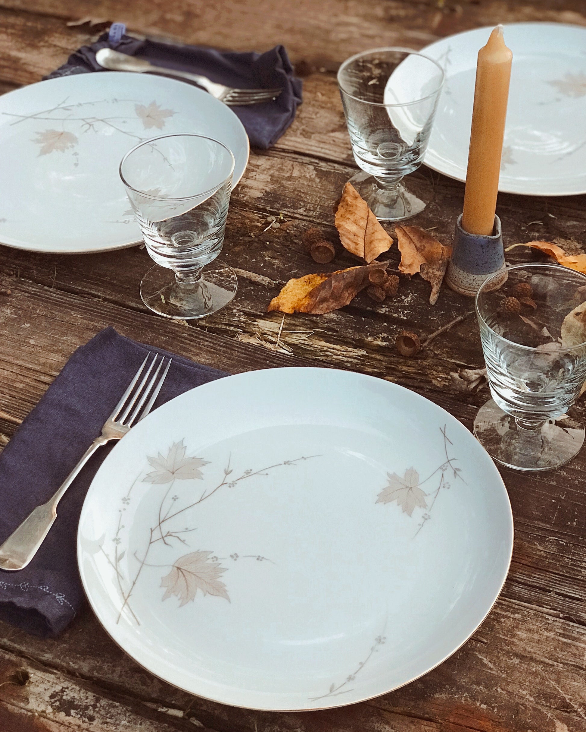 Set of plates, glasses, and cutlery on a wooden table with autumn leaves.