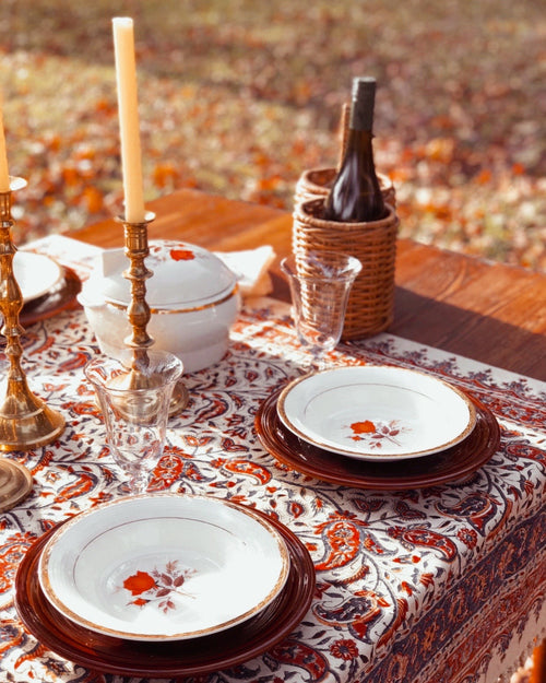 Dining table set with plates, glasses, and a bottle on a patterned tablecloth outdoors.