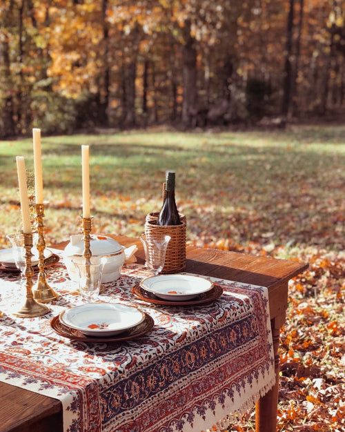 Dining table set with plates, glasses, and candles outdoors in a forest setting.