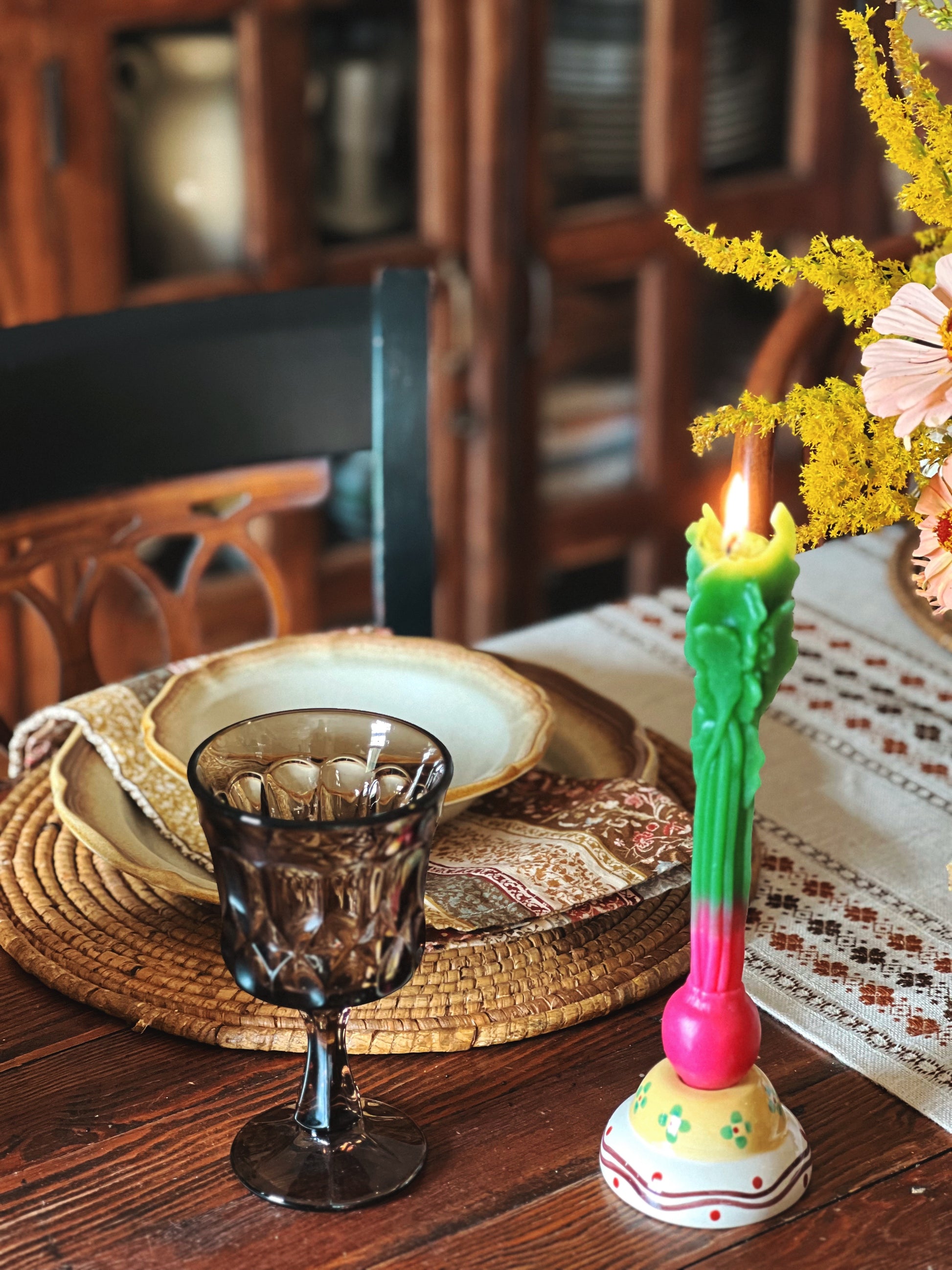 Decorative table setting with a colorful candle, glass goblet, and floral arrangement on a wooden table.