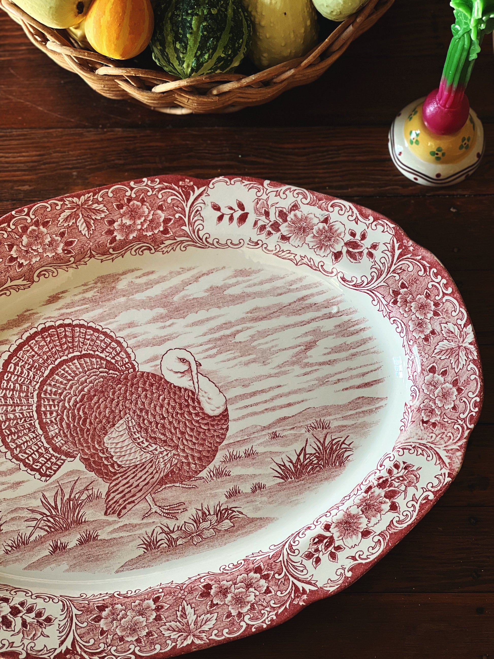 Decorative platter with a red turkey design on a wooden surface with a basket of fruits.