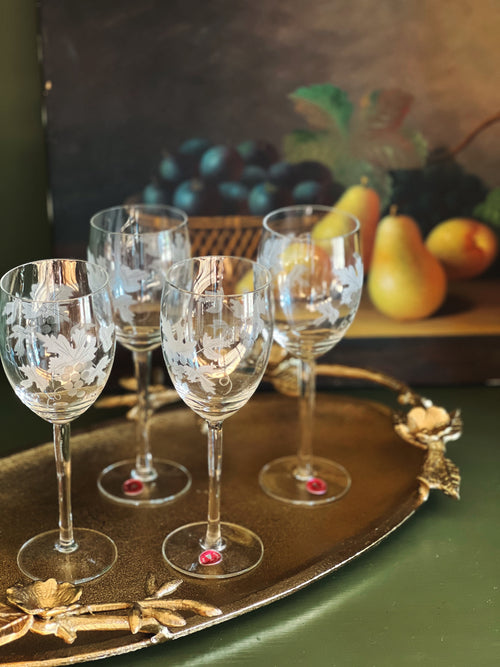 Set of four etched wine glasses on a decorative tray with fruit in the background