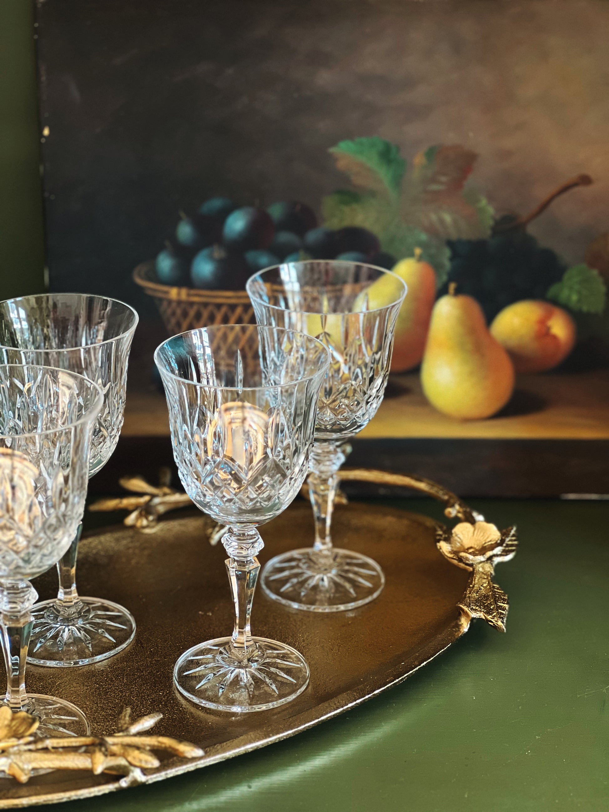 Set of crystal glasses on a decorative tray with a fruit still life painting in the background.