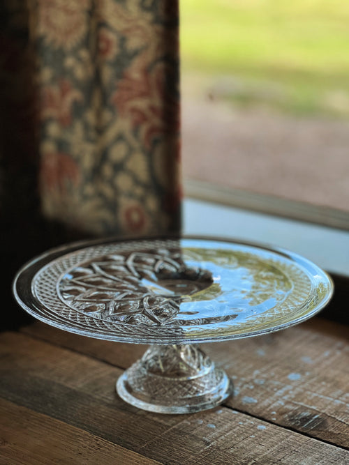 Decorative glass cake stand on a wooden surface with a blurred floral-patterned curtain in the background.