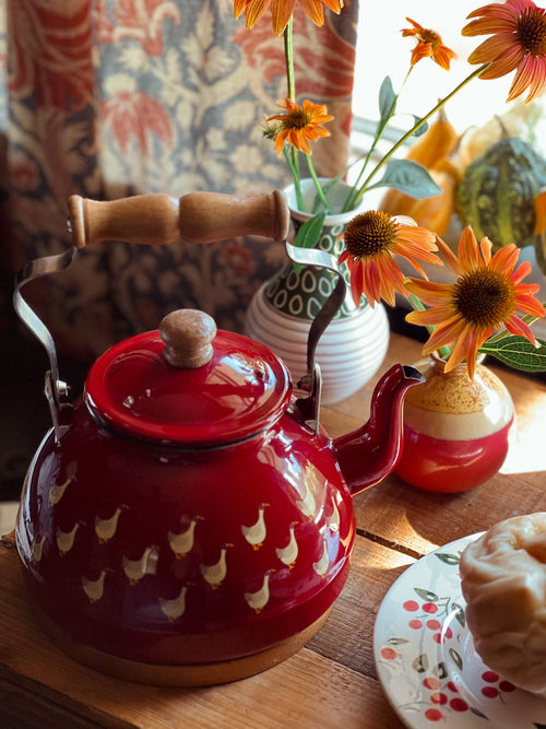 Red teapot with a wooden handle on a table with flowers and a plate.