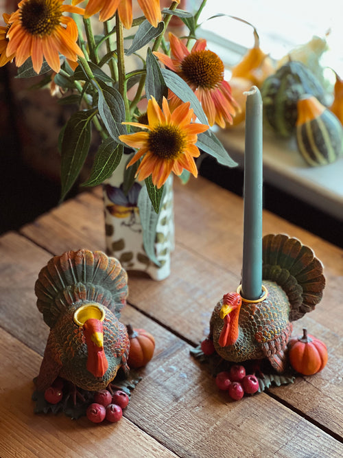 Two turkey-shaped candle holders on a wooden surface with flowers and pumpkins in the background.