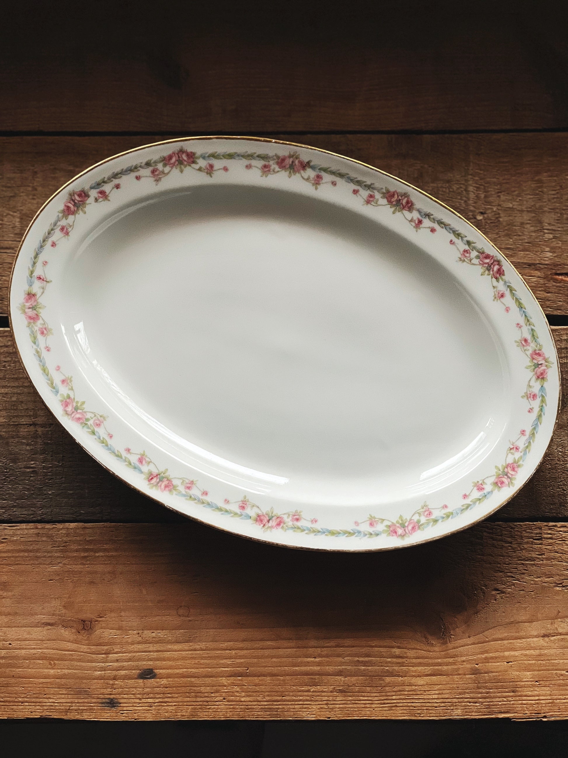 An oval-shaped porcelain serving platter with pink roses and blue/green laurel garland design, displayed on a wooden surface.