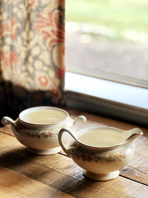 Two ceramic creamers and sugar bowl on a wooden surface with a floral curtain and window in the background.