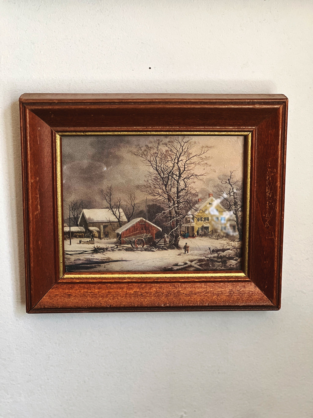 Framed painting of a winter scene with a house and barn on a white wall.