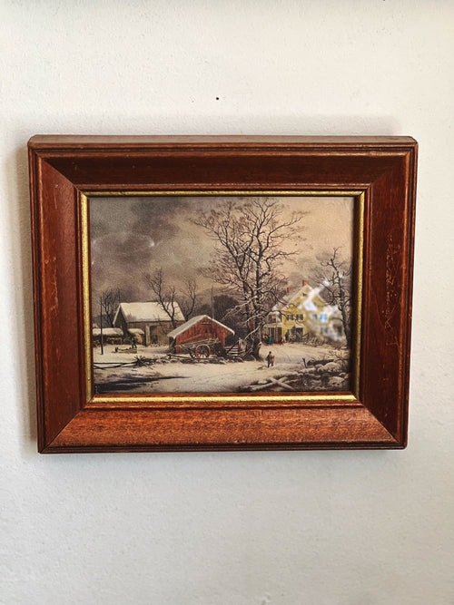 Framed painting of a winter scene with a house and barn on a white wall.