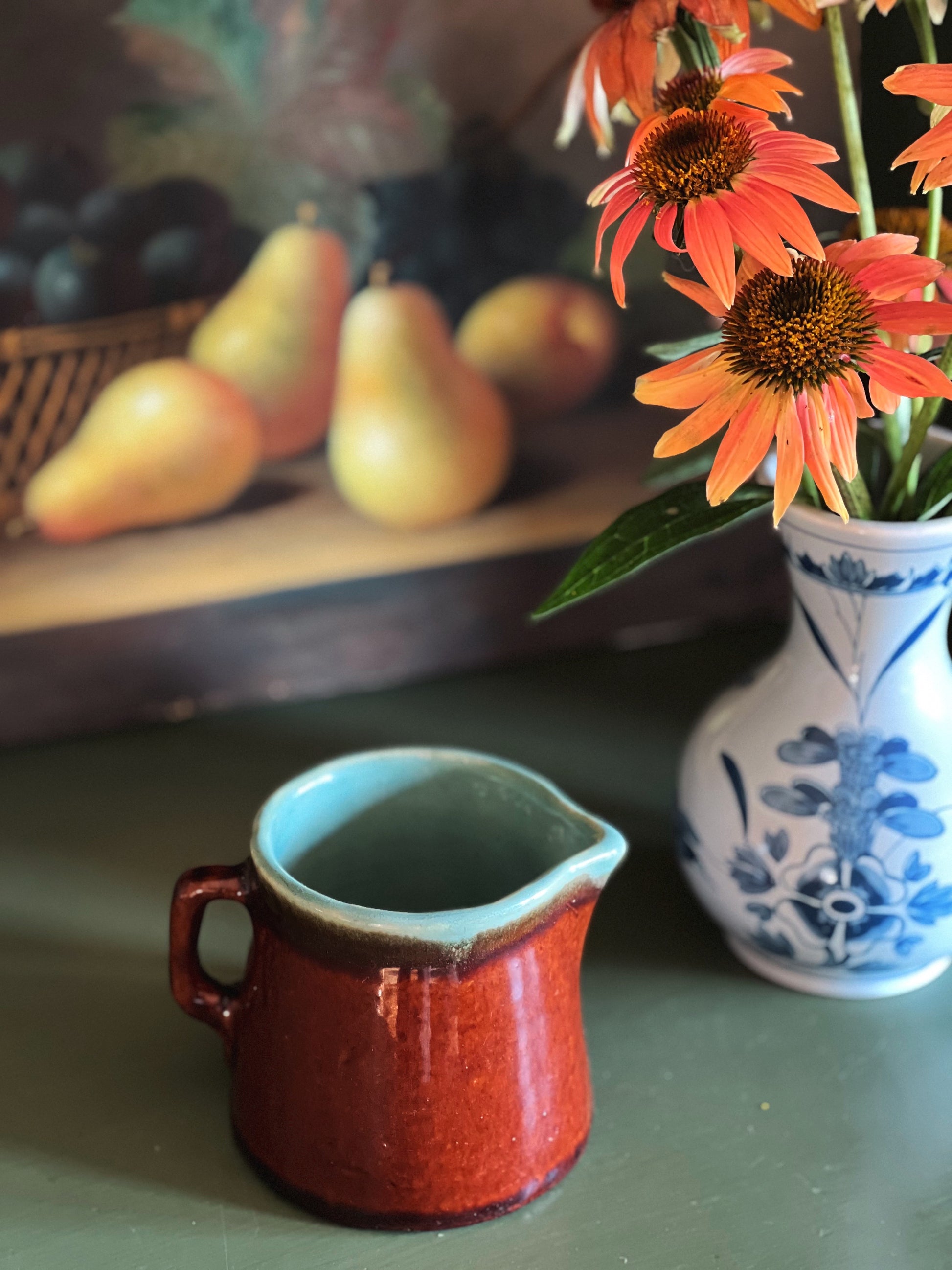 Red ceramic pitcher on a table with a vase of flowers and pears in the background
