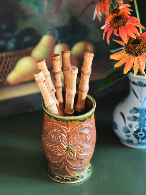 Ceramic holder with bamboo sticks on a table with flowers and fruit in the background