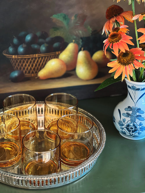 Tray of glasses with amber liquid, basket of fruit, and vase with flowers on a table.