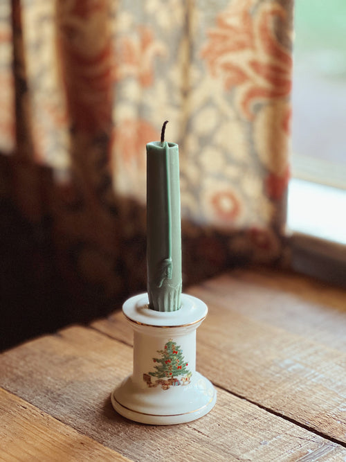 Decorative candle in a white holder with Christmas design on a wooden surface, floral curtains in the background.